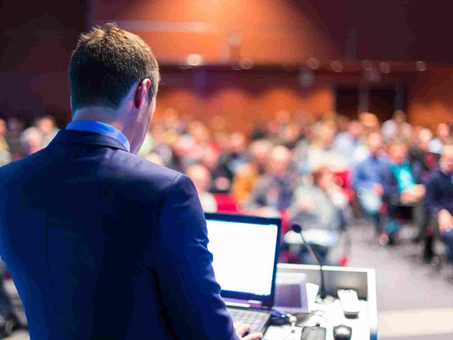A person with a laptop standing on a stage in front of many people (640x480)